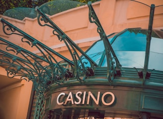 The image displays the entrance to a casino, characterized by detailed and ornate architectural elements with green metal structures and a sign that reads 'CASINO'. The background includes a glass dome and a peach-colored wall.