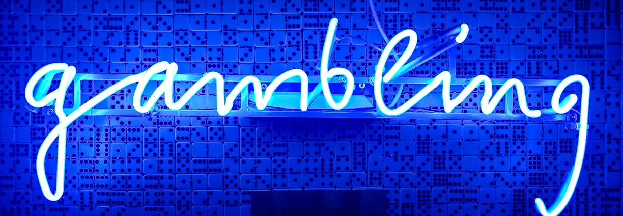 A bright blue neon sign spells out the word 'gambling' in cursive script. The background features a textured wall covered in domino tiles, also illuminated by the blue light, creating a vivid and visually striking scene.