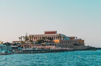A seaside view of a large building labeled as a casino, situated on a rocky shore. The building is surrounded by palm trees and other smaller structures. The calm sea water reflects the sky, and there are several closed umbrellas lined along the waterfront.