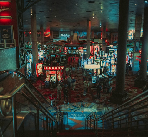 A bustling casino floor filled with groups of slot machines and people walking around. The atmosphere is vibrant with neon lights and illuminated signs. The interior has a ceiling supported by large pillars and there are stairs leading down to the gaming area.